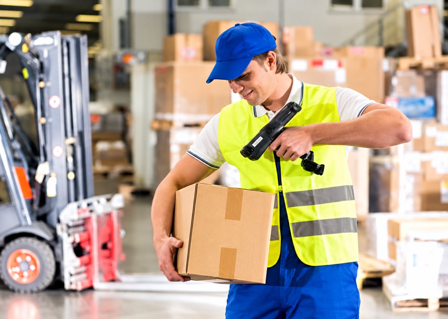 Un employé en casquette bleue et gilet jaune tient un carton et le scanne avec un lecteur portable dans un entrepôt, près d’un chariot élévateur.