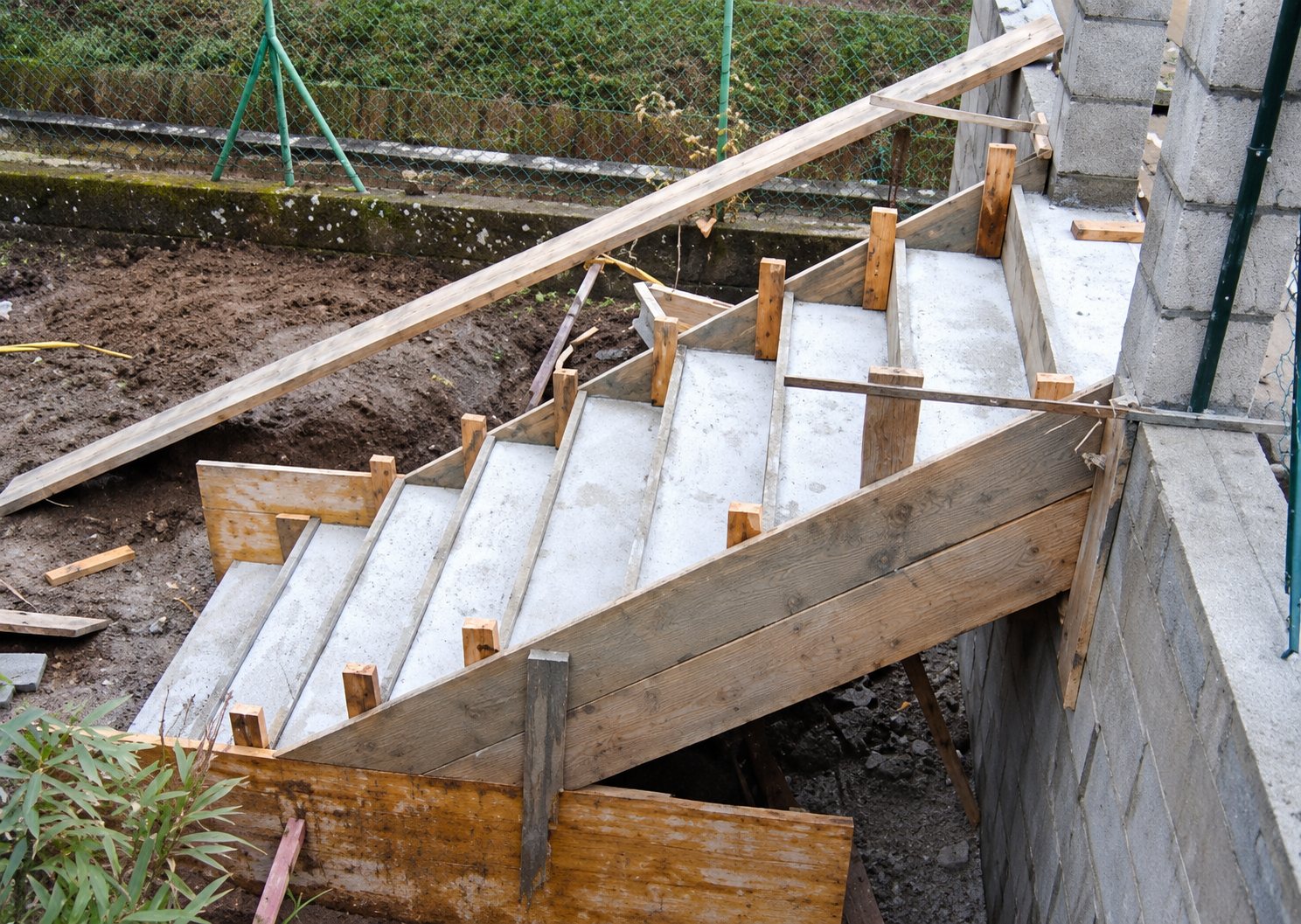 Un escalier en béton fraîchement coulé, encore maintenu par son coffrage en bois, sur un chantier en extérieur près d’un mur.