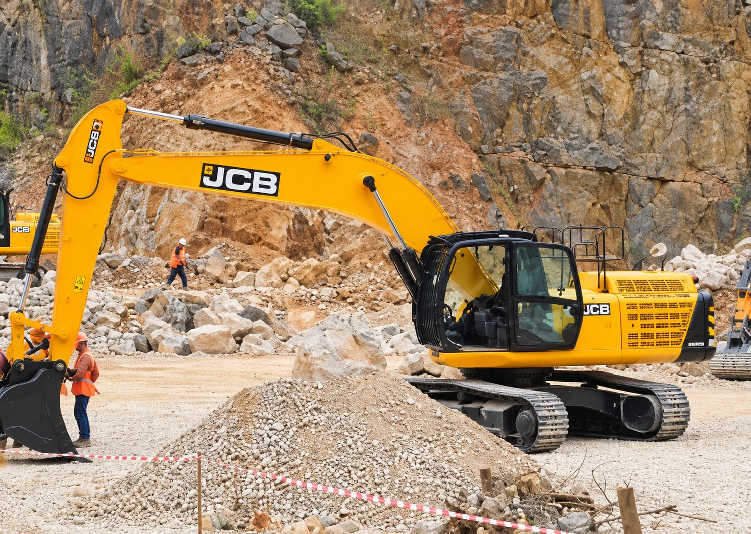 Une grande pelleteuse JCB orange et jaune travaille dans une carrière, devant une paroi rocheuse et un tas de gravats, avec des ouvriers à proximité.